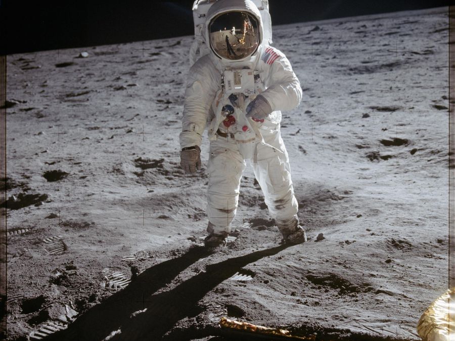 An astronaut standing on the gray lunar surface, the photographer reflected in his visor